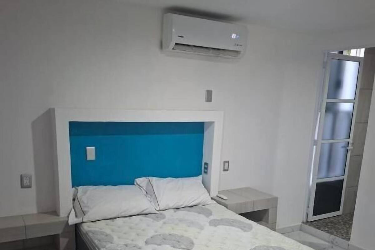Un dormitorio con cama, cabecera azul, pared blanca, unidad de aire acondicionado y ventana.