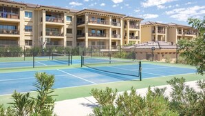 Ein Tennisplatz mit Netz, umgeben von einem Zaun, neben einem mehrstöckigen Gebäude mit Balkonen.