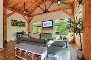 Ein geräumiger Wohnbereich mit Holzbalkendecke, einem großen Sofa, einem Fernseher und Blick ins Grüne.