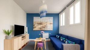 A modern living room with a blue sofa, a wooden TV stand, a small table, and a large abstract painting on the wall. - Blue House - Parking - Downtown (Reims)