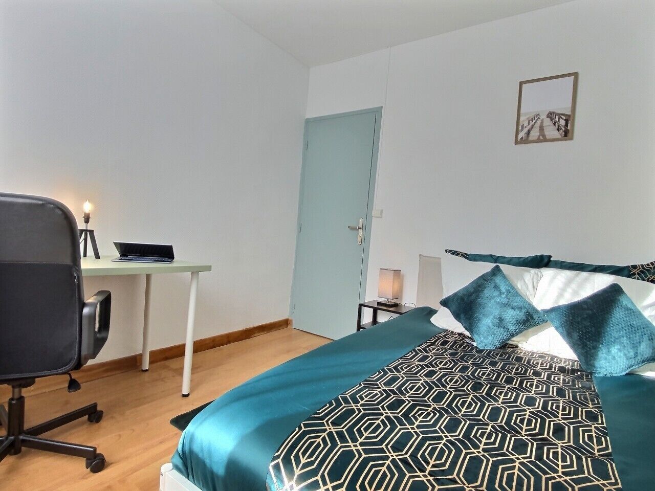 A bedroom with a bed, a desk, a chair, a door, and a wall-mounted picture.