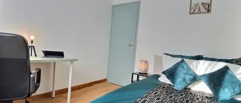 A bedroom with a bed, a desk, a chair, a door, and a wall-mounted picture.