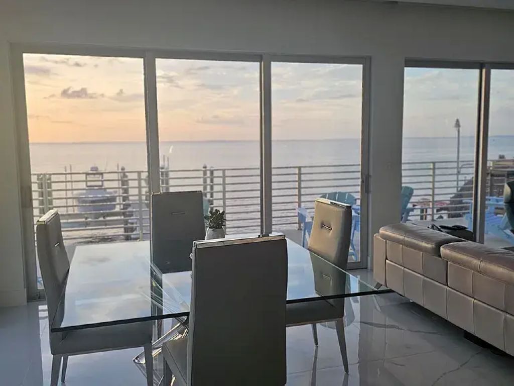 Una sala de estar moderna con una mesa de vidrio, sofá de cuero y grandes ventanas con vista a un puerto deportivo.