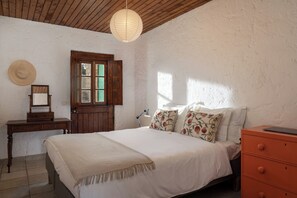 Une chambre avec un plafond en bois, un lit agrĂ©mentĂ© dâoreillers Ă motifs floraux, une commode en bois et un bureau avec un miroir.