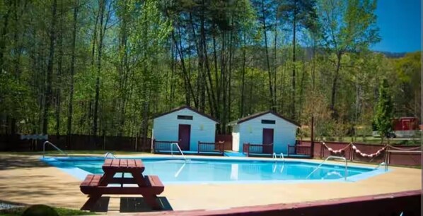 A swimming pool with a picnic table in the foreground, surrounded by trees and two small buildings in the background.