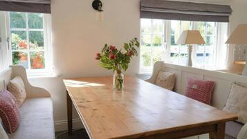 A room with a wooden table, a vase with flowers, a lamp, and a view of greenery through the windows.
