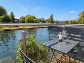 A balcony with a table and chairs overlooking a canal, with a bottle of wine and a glass on the table. - Island Cottage Rod Eyot (Henley-on-Thames)