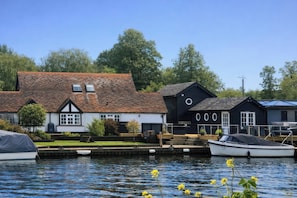 A riverside building with a docked boat, a small garden, and a clear sky. - Island Cottage Rod Eyot (Henley-on-Thames)