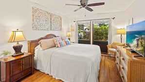 A bedroom with a bed, bedside tables, a ceiling fan, a large window with a view, and a television. - Walk to Poipu Beach, Grand Hyatt & Brennecke's/Poipu Kai Charming Condo (Koloa)