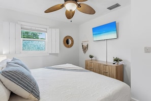 Ein Schlafzimmer mit einem Bett, einem Deckenventilator, einem Fenster mit Jalousien, einem an der Wand montierten Fernseher und einer Holzkommode.