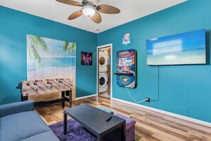 Ein Zimmer mit Wandgemälde zum Strandthema, Deckenventilator, Waschmaschine, Fernseher und einem Kicker.