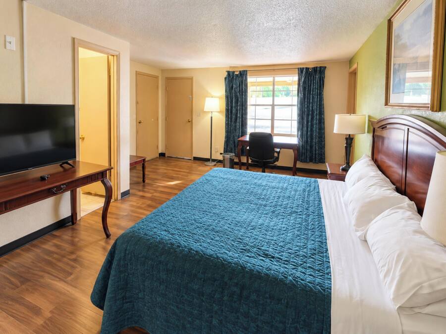 Ein Hotelzimmer mit einem Bett, einem Schreibtisch mit Fernseher, einem Sessel und einem Fenster mit Vorhängen.