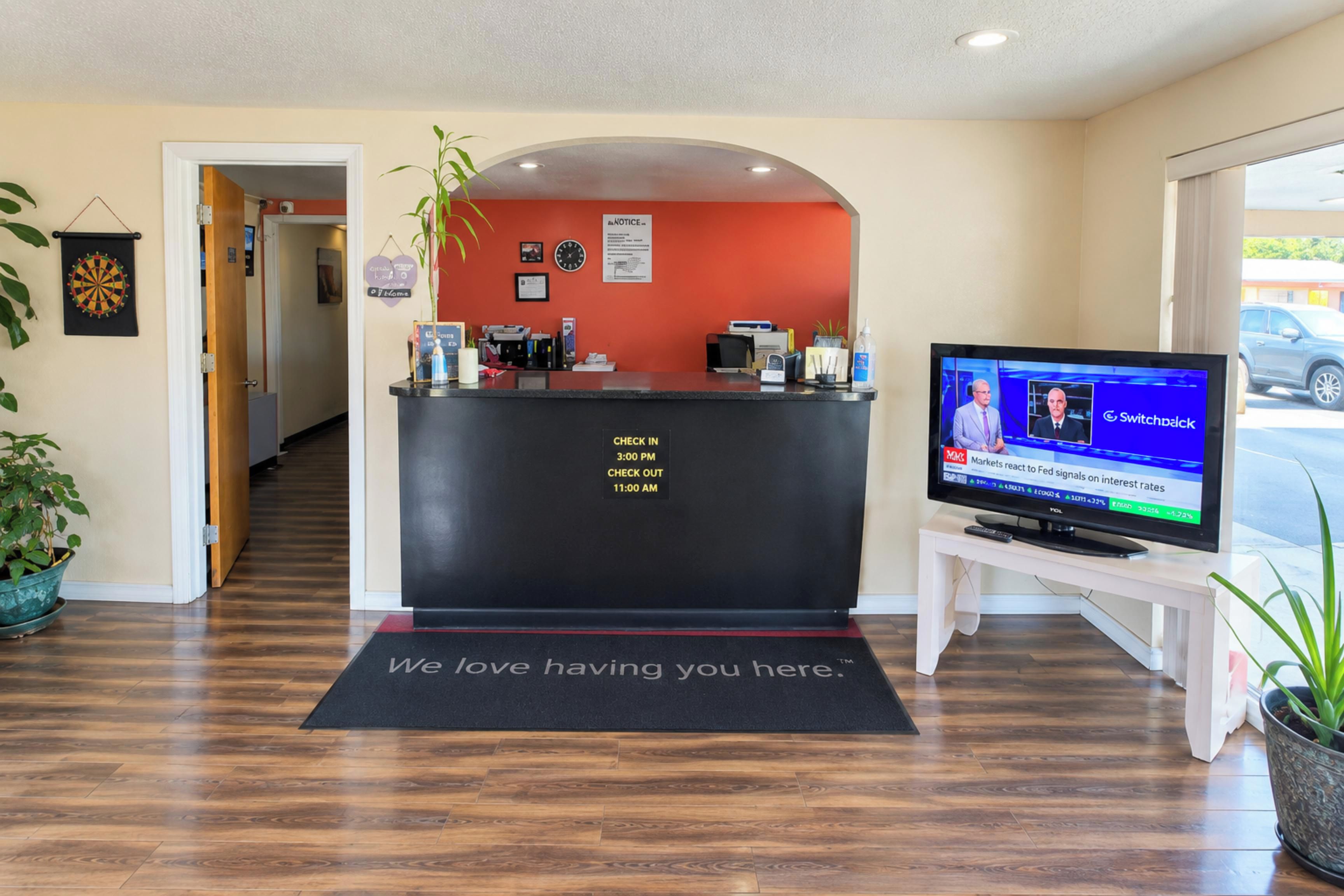 Zona de recepción con un mostrador negro, un televisor mostrando un noticiero y una alfombra de bienvenida.