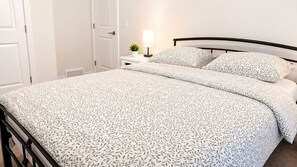 A neatly made bed with a patterned bedspread, a bedside table with a lamp, and a potted plant. - Heartland Stays - Brand New 3 Bed Room Cozy Home with AC, Garage, BBQ (Cochrane)