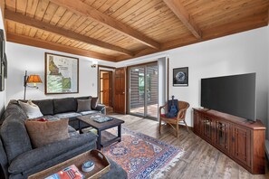 A living room with a sofa, coffee table, TV, wooden cabinet, and a large window. - Pilgrim Cove Basecamp: 4BR, 3BA mountain-view retreat in McCall. (McCall)