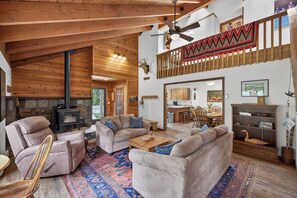A cozy living room with a fireplace, wooden ceiling, and a high ceiling fan. - Pilgrim Cove Basecamp: 4BR, 3BA mountain-view retreat in McCall. (McCall)