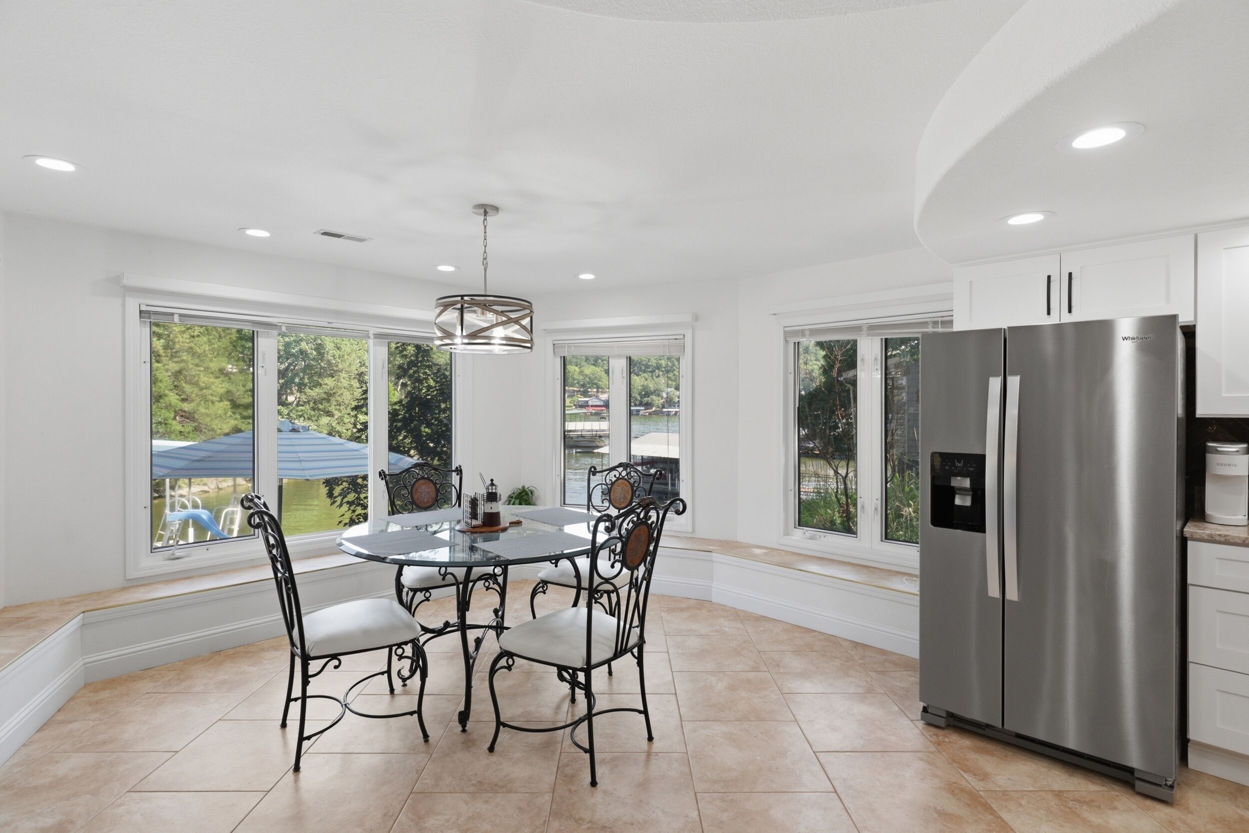 A modern kitchen with a stainless steel refrigerator, a dining table with four chairs, and large windows offering a view of the outdoors.