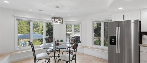 A modern kitchen with a stainless steel refrigerator, a dining table with four chairs, and large windows offering a view of the outdoors.