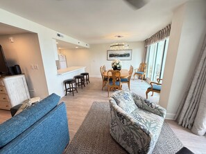 Ein modernes Wohnzimmer mit Sofa, Essbereich mit Tisch und Stühlen, Küche mit Kühlschrank und einem großen Fenster.