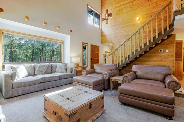 A spacious living room with a large window, a wooden staircase, and leather sofas. - 20 Camas - Sunriver Charm, Hot Tub, Fireplace, AC, 10 SHARC Passes (Sunriver)