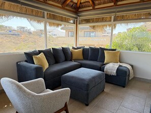 A modern living room with a sectional sofa, ottoman, and armchair, large windows, and a thatched roof ceiling. - Private Home—Ocean View with Pool and Fire Pit (Perfect for Vacations) (BCS)