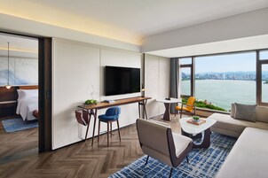 Una habitación de hotel moderna con un ventanal grande, televisor de pantalla plana, sofá, mesa de comedor y una pequeña cocina.