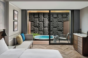 Habitación de hotel moderna con una cama grande, un sofá, una mesa de comedor, una pequeña cocina y vista a una piscina a través de una puerta de vidrio.