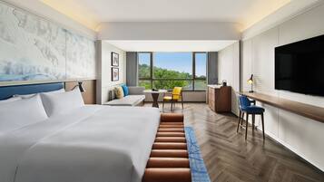 Habitación de hotel con una cama grande, un escritorio, una silla, un televisor y vistas al exterior con zonas verdes.
