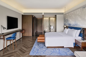 Habitación de hotel con cama, escritorio, silla, televisión y baño con espejo y retrete.