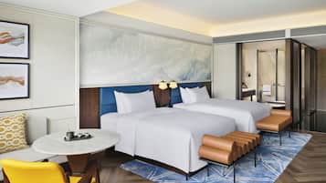 A hotel room with two beds, a round table, a chair, a bathroom with a mirror, and artwork on the wall.