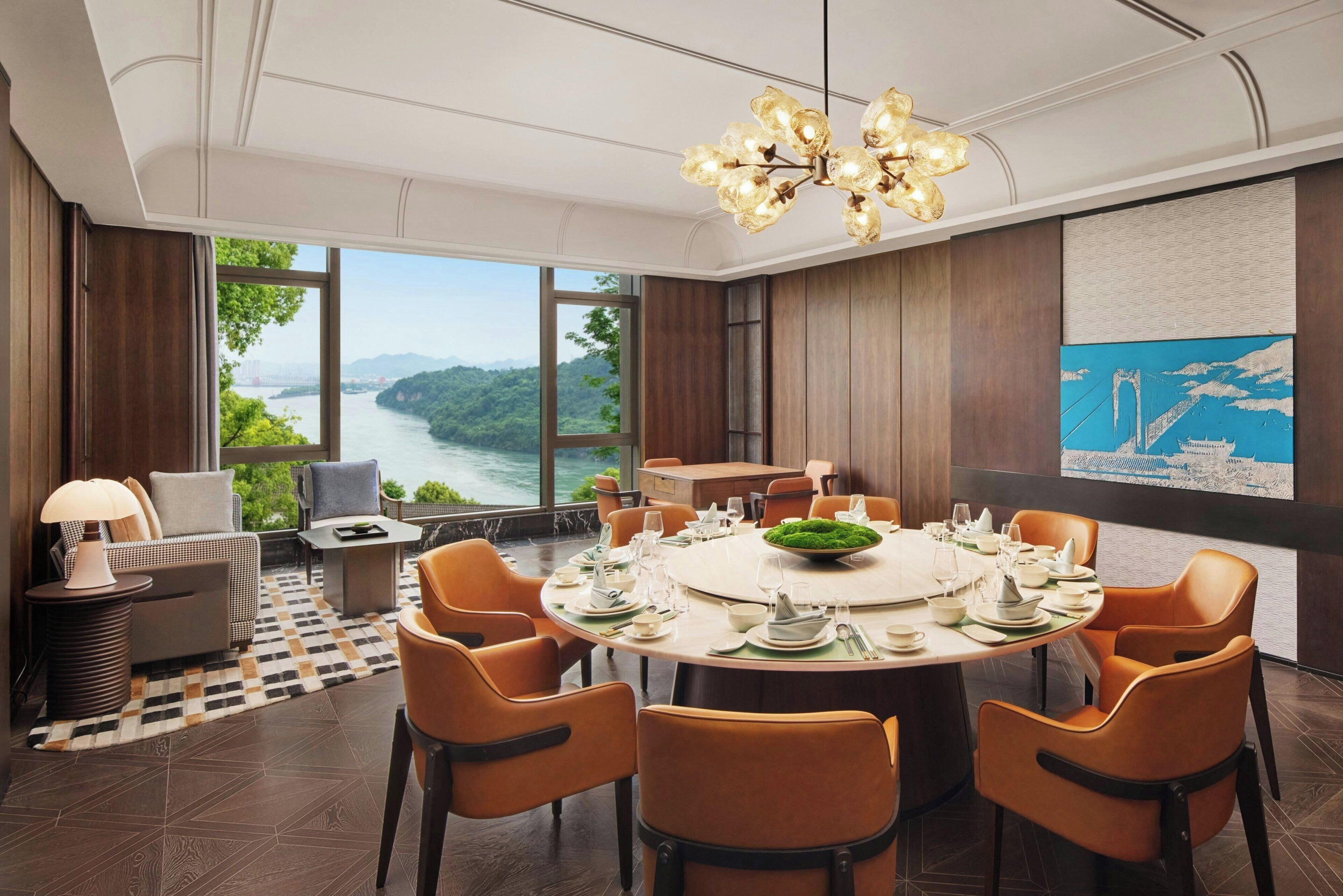 Un comedor con una mesa redonda dispuesta para seis personas, rodeada de sillas color naranja, con vista a un río y montañas a través de grandes ventanales.