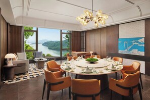 A dining area with a round table set for six, surrounded by orange chairs, with a view of a river and mountains through large windows.