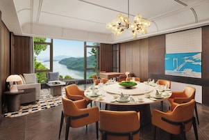 Un comedor con una mesa redonda dispuesta para seis personas, rodeada de sillas color naranja, con vista a un río y montañas a través de grandes ventanales.