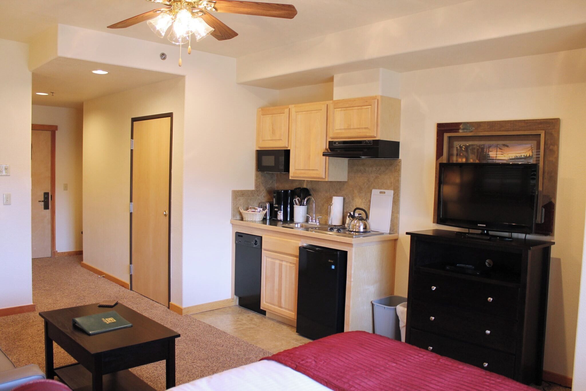 A compact hotel room with a kitchenette, a bed, a TV, and a ceiling fan.