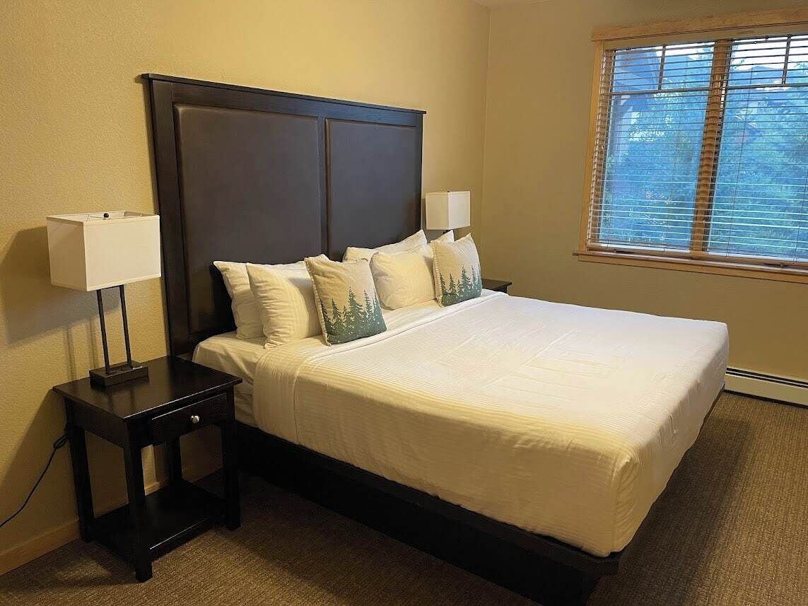 A neatly made bed with a dark headboard, two bedside tables with lamps, and a window with blinds.