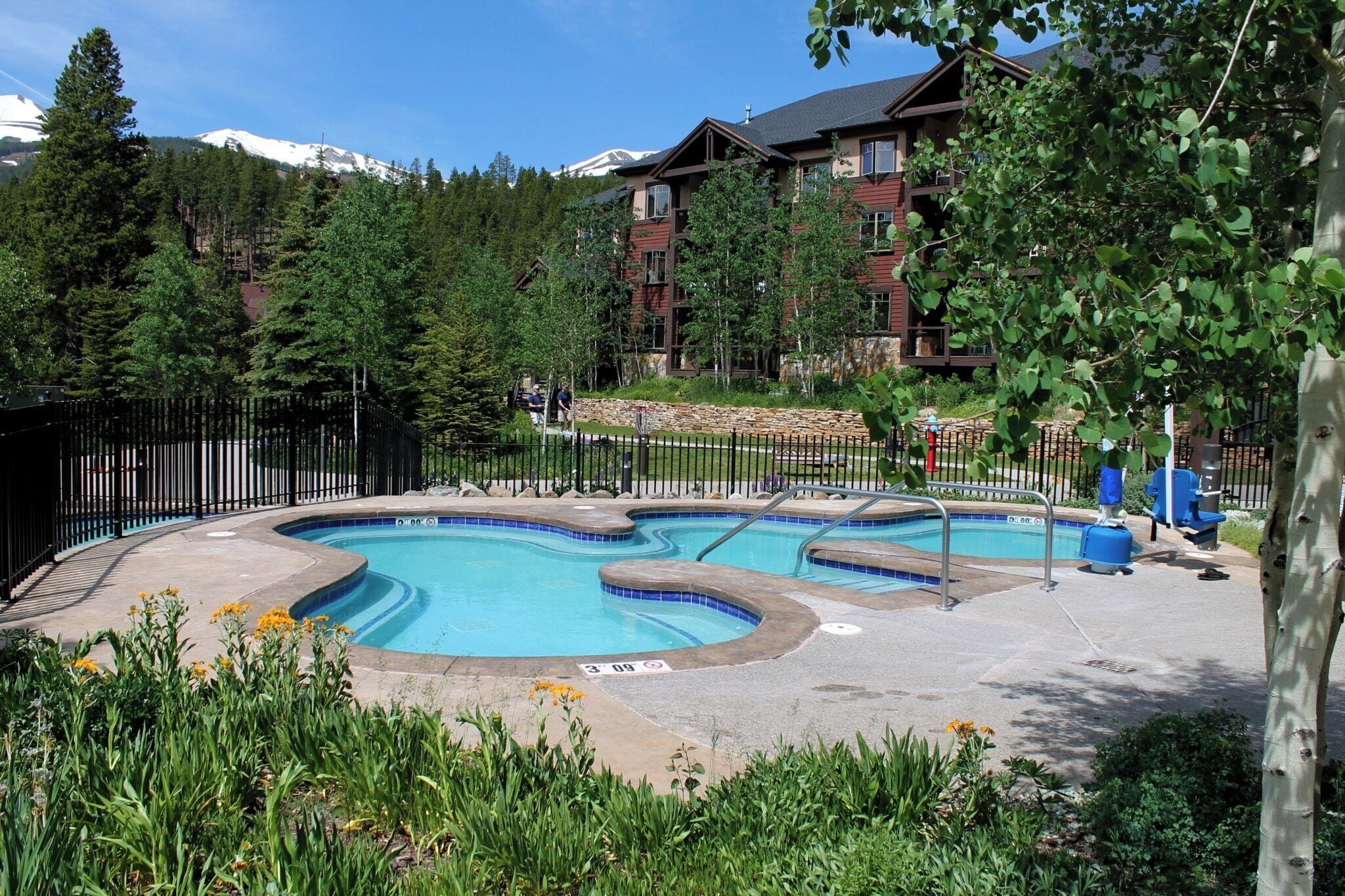 A swimming pool with a curved design, surrounded by a black fence, with a building in the background and a mountainous landscape.