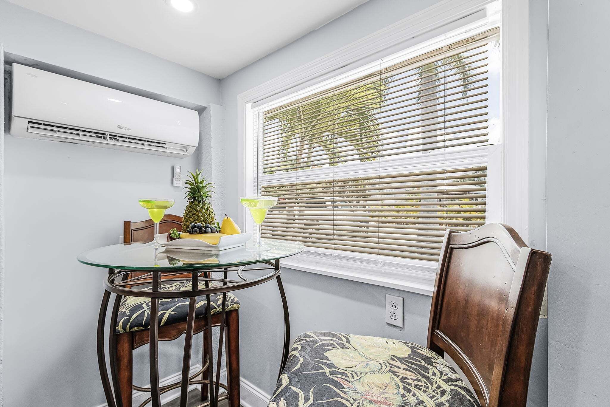 A dining area with a glass table, chairs, a small plant, and a window with blinds.