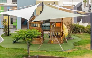 Ein Kinderspielbereich mit Schaukeln, einer Rutsche und einer Kletterlandschaft, umgeben von einer Rasenfläche und einem Gebäude im Hintergrund.