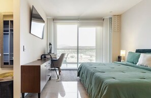 Un dormitorio con cama, escritorio, silla, televisor y un ventanal con vistas.