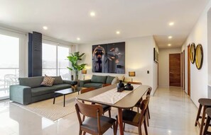 A modern living room with a sofa, coffee table, dining table, and chairs. There is a large window with a view, a plant, and artwork on the wall. - Cozy and Modern Apartment. (Juriquilla)