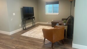Un salon avec un téléviseur posé sur un meuble, un fauteuil en cuir marron, un tapis blanc et une fenêtre avec des stores.