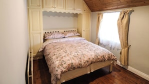 A bedroom with a wooden ceiling, a bed with floral bedding, a wardrobe, and a window with curtains. - Gullane East (Killarney)