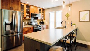 A kitchen with stainless steel appliances, wooden cabinets, a large island with bar stools, and a pendant light. - The Northport Sapling | In-Town Northport | Walk to Bay, Shops + Dining (Northport)