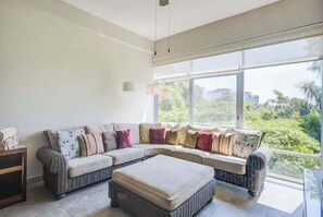 Un salon moderne avec un grand canapé d’angle, une petite table et une grande fenêtre donnant sur un paysage verdoyant.