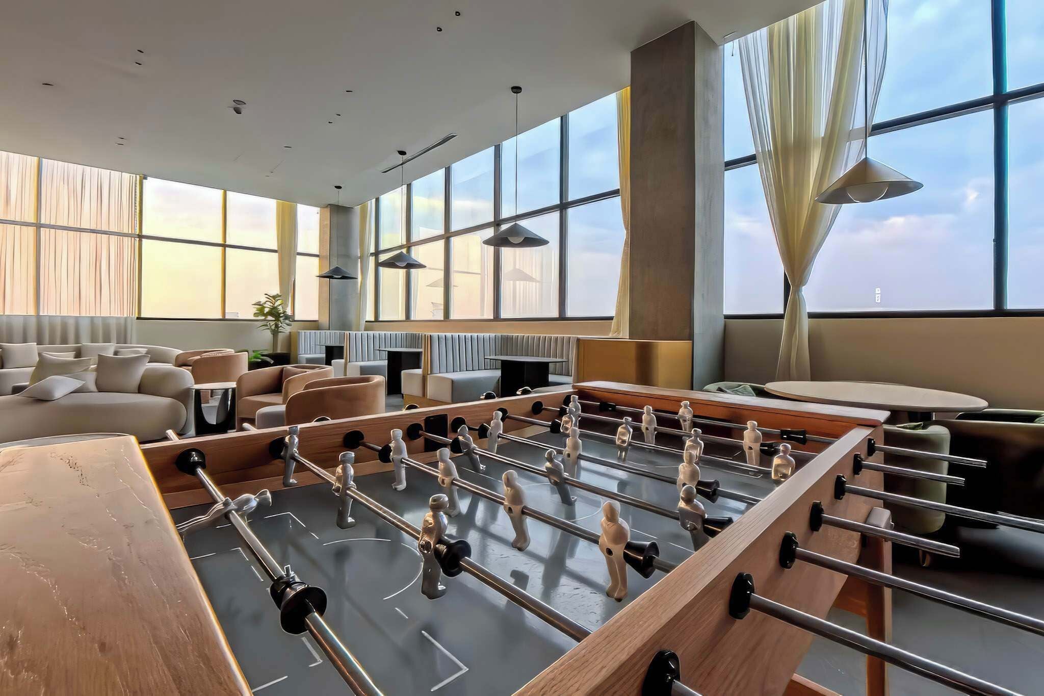 A modern hotel room with a foosball table, a sofa, a dining table, and large windows.