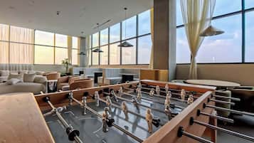 A modern hotel room with a foosball table, a sofa, a dining table, and large windows.
