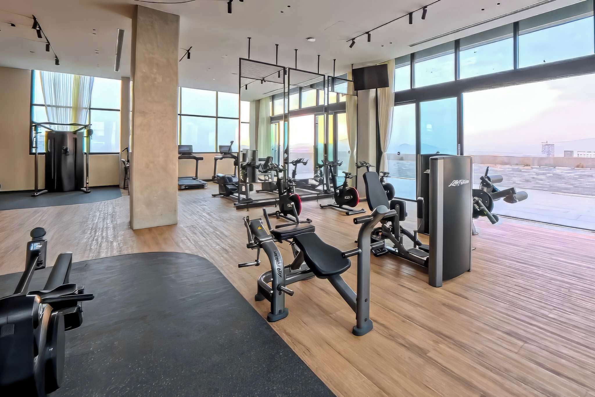 A modern gym with various exercise equipment, large windows, and a view of the sky.
