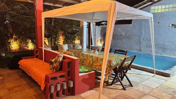 A covered outdoor seating area with a table and chairs, a pool in the background, and a red structure with a bench. - Beach house in Alemão offering comfort near the sea. House with pool, near the sea. (Caponga)