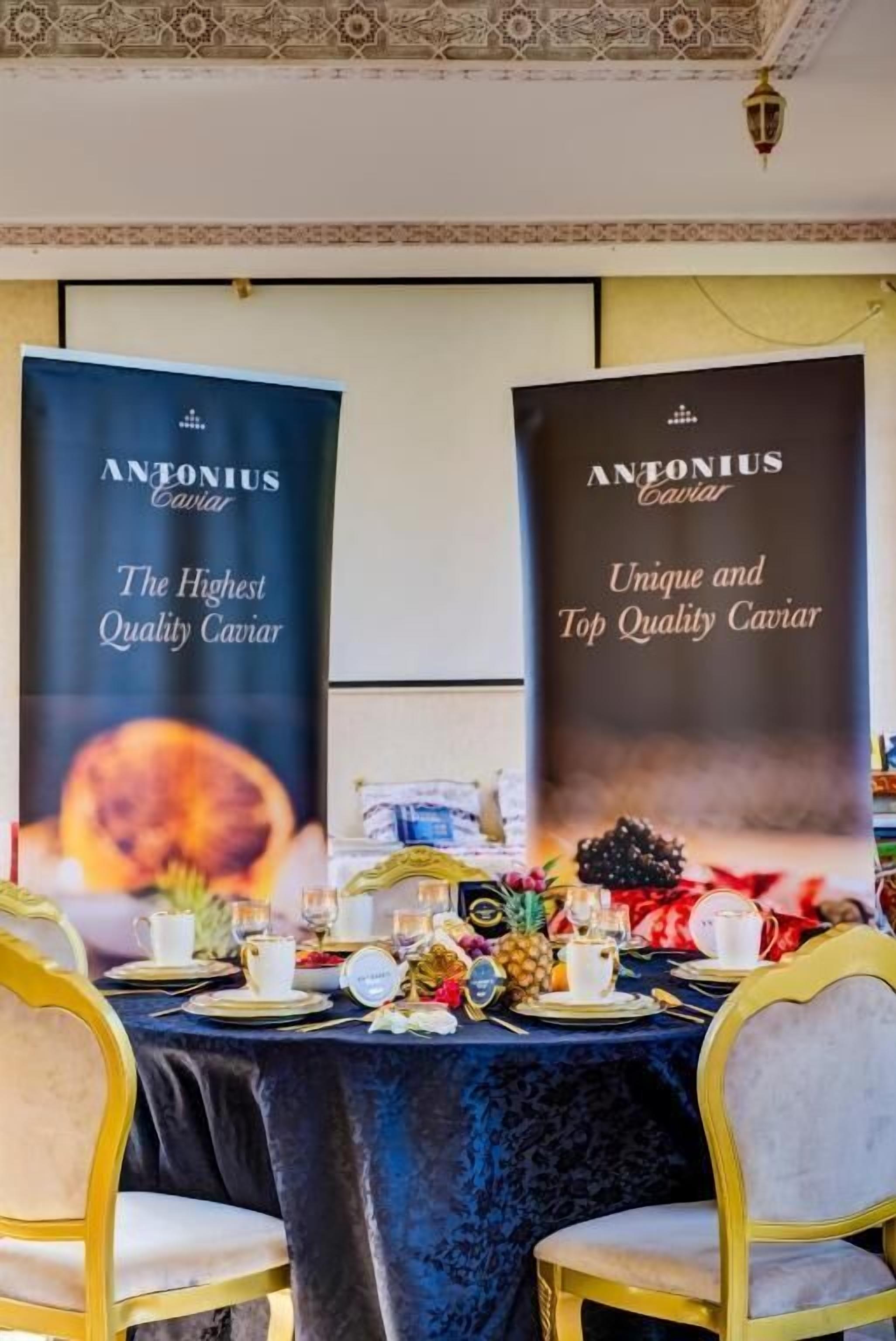 Uma mesa posta para refeição, com toalha de mesa preta, cadeiras douradas e um banner publicando 'ANTONIUS Caviar'.