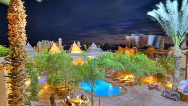 A pool area with lounge chairs and umbrellas, surrounded by palm trees and a building with multiple domes. - Riad Jnane Ines (Machraa El Ain)